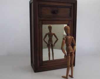 May include: A wooden cabinet with a single drawer and a mirror door. A wooden articulated doll stands in front of the cabinet, looking at its reflection in the mirror.