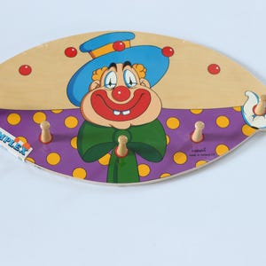 Op de afbeelding: Een houten kapstok in de vorm van een ovaal met een cartoonclownontwerp. De clown draagt een paars-geel gestippeld shirt, een groene strik en een blauwe hoed met gele bies. De kapstok heeft vijf haken om spullen aan op te hangen.