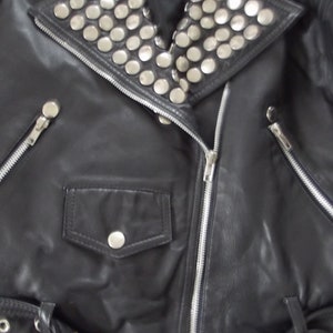 May include: Black leather jacket with silver metal studs on the collar. The jacket features a zippered front, a small pocket with a button, and a belt with a buckle. The jacket is a classic biker style.