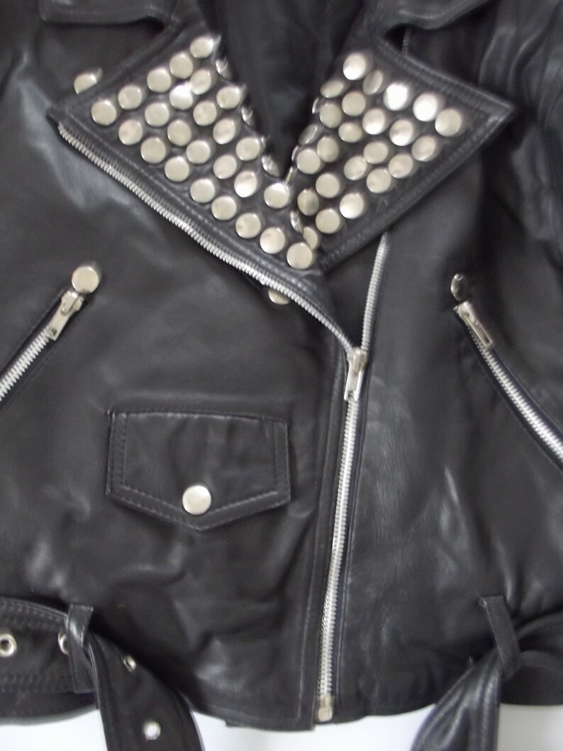 May include: Black leather jacket with silver metal studs on the collar. The jacket features a zippered front, a small pocket with a button, and a belt with a buckle. The jacket is a classic biker style.