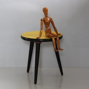 May include: A small yellow and black round table with three black legs. A wooden artist's mannequin is sitting on the table.