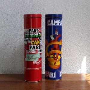 May include: Two cylindrical metal tins featuring Campari branding. The left tin is red with repeated white and yellow Campari text and a small logo. The right tin is blue, displaying the Campari logo, a figure, and a large orange peel graphic.