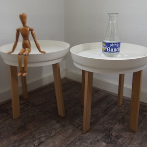 Op de afbeelding: Twee witte ronde bijzettafels met houten poten. Op de ene tafel zit een houten figuur. Op de andere tafel staat een heldere glazen fles met een blauw en wit etiket waarop "Gancia" staat.