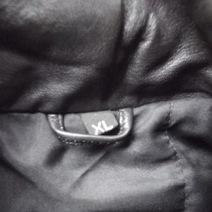 May include: Close-up of a black leather jacket interior. A small black tag with the white letters "XL" is visible. The jacket's lining is black.