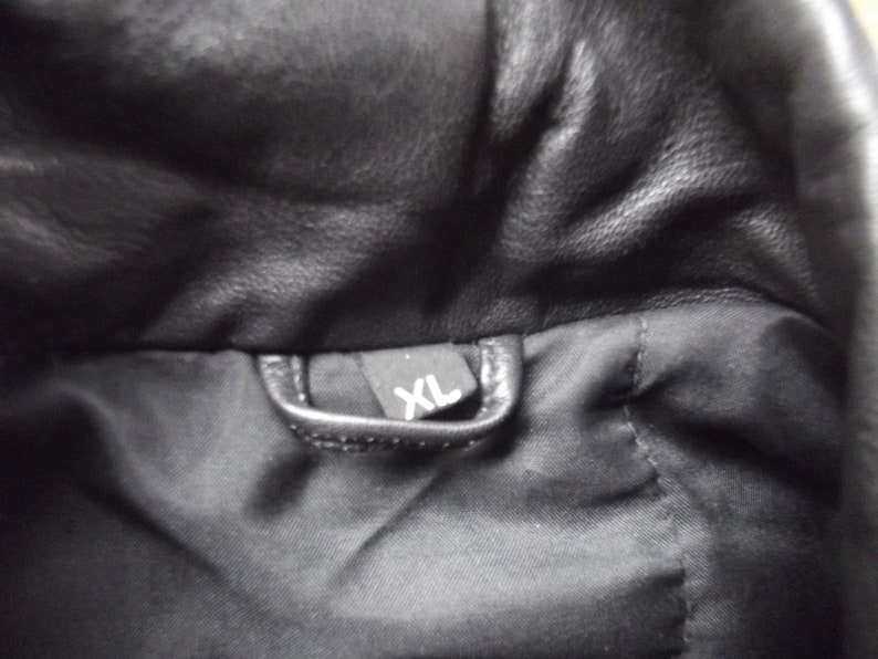 May include: Close-up of a black leather jacket interior. A small black tag with the white letters "XL" is visible. The jacket's lining is black.