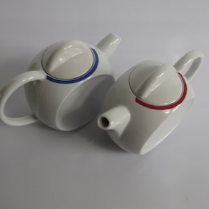 Może przedstawiać: Dwa białe ceramiczne czajniki z niebieskimi i czerwonymi akcentami. Czajnik po lewej stronie ma niebieski akcent wokół pokrywki, a czajnik po prawej stronie ma czerwony akcent wokół pokrywki.
