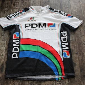 May include: White and black cycling jersey with the text "PDM CHROME CASSETTES". The jersey features a colorful design with red, green, and blue stripes. The sleeves and sides have the "PDM" logo.