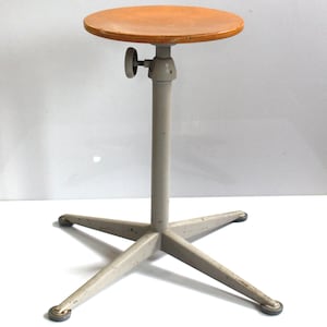 May include: A height-adjustable stool featuring a round, light brown wooden seat mounted on a central gray metal column. The base consists of four gray metal legs arranged in a cross pattern, each ending in a small, circular foot pad.