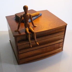 Puede incluir: Un organizador de escritorio de madera con tres cajones horizontales y tiradores integrados. Un pequeño maniquí articulado de madera está sentado sobre la superficie superior junto a dos libros apilados con cubiertas marrón y azul.