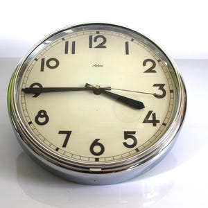 Puede incluir: Un reloj de pared redondo con marco de cromo pulido y esfera de color crema. El reloj presenta números arábigos negros del uno al doce, manecillas negras para las horas y los minutos, y la marca "Melns" impresa en negro bajo el doce.