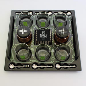 May include: A black square serving tray featuring a printed green circuit board pattern. The tray holds six clear glass cups, two black cylindrical containers with white cross logos, and six small silver spoons with resistor-like handles.