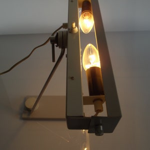 K&ouml;nnte beinhalten: Eine wei&szlig;e Metall-Tischlampe mit zwei Gl&uuml;hbirnen. Die Lampe hat einen langen Arm und einen rechteckigen Sockel.