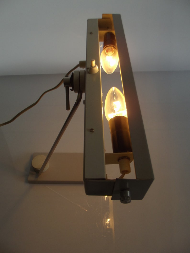 K&ouml;nnte beinhalten: Eine wei&szlig;e Metall-Tischlampe mit zwei Gl&uuml;hbirnen. Die Lampe hat einen langen Arm und einen rechteckigen Sockel.