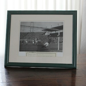 May include: Framed black and white photograph of a soccer game in progress. The image shows a goalkeeper diving to save a shot on goal. The frame is dark green.