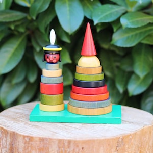 Puede incluir: Un juguete de apilar de madera con dos torres. Una torre es roja, verde, amarilla y negra. La otra torre tiene una cara roja y negra con una pluma en la parte superior. Ambas torres están sobre una base verde.