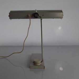 K&ouml;nnte beinhalten: Eine silberne Schreibtischlampe mit einem langen, rechteckigen Schirm und einer einzelnen Gl&uuml;hbirne. Die Lampe hat einen Metallfu&szlig; und ein Kabel mit einem Stecker.