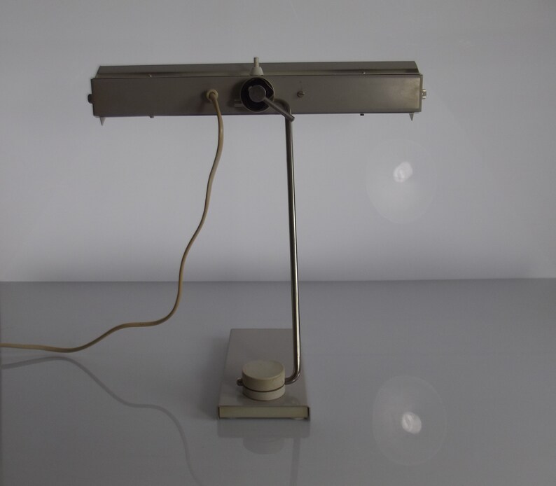 K&ouml;nnte beinhalten: Eine silberne Schreibtischlampe mit einem langen, rechteckigen Schirm und einer einzelnen Gl&uuml;hbirne. Die Lampe hat einen Metallfu&szlig; und ein Kabel mit einem Stecker.
