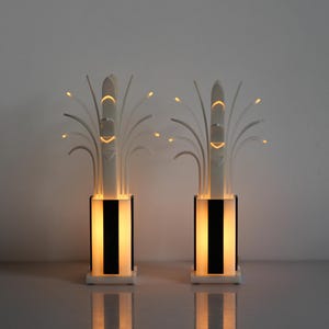 May include: Two white table lamps with black accents and a unique design resembling a stylized plant with multiple branches. The lamps are illuminated with warm white light.