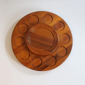 Op de afbeelding: Een ronde houten serveerschaal met een centraal verhoogd gedeelte en tien cirkelvormige uitsparingen. De schaal is gemaakt van warm getint hout, waarschijnlijk walnoot of teak, en heeft een gladde, gepolijste afwerking. Het ontwerp suggereert dat het bedoeld is voor het serveren van kleine items.