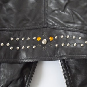 May include: Black leather jacket with silver studs and orange gemstones. The jacket features a decorative studded band across the upper back. A silver zipper is visible on the front.