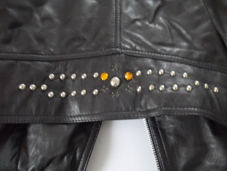 May include: Black leather jacket with silver studs and orange gemstones. The jacket features a decorative studded band across the upper back. A silver zipper is visible on the front.