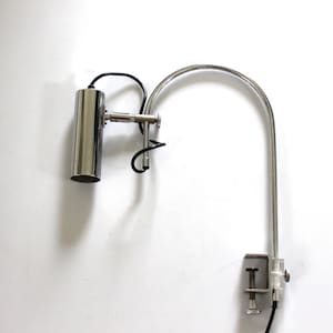 May include: A silver-colored, adjustable desk lamp with a cylindrical head and a curved arm. The lamp has a clamp base for secure attachment and a black power cord. The design is minimalist and modern, suitable for various settings.