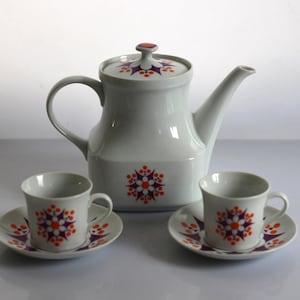 Może przedstawiać: Biały ceramiczny czajnik i dwie pasujące filiżanki do herbaty z podstawkami. Czajnik ma geometryczny wzór z pomarańczowymi i fioletowymi kwiatami. Filiżanki i podstawki mają podobny wzór.