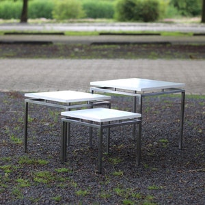 Op de afbeelding: Drie witte nesttafels met zwarte metalen frames. De tafels zijn in een verspringende opstelling op een grind oppervlak geplaatst.