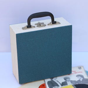 Puede incluir: Un maletín de discos de vinilo vintage con frente verde azulado y laterales blanquecinos, con asa negra y herrajes plateados. Se ven varios discos de vinilo, lo que sugiere un tema de almacenamiento de música.