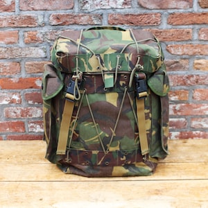 May include: A camouflage pattern backpack featuring a top flap with elastic cord lacing, two side pockets, and two vertical tan nylon straps with black plastic buckles. The main compartment is secured with a drawstring and a top lid closure system.