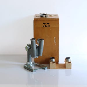 May include: A vintage microscope with a grey metal body and two black eyepieces. It is sitting on a white surface next to a wooden box with the number 55 on the front. The box has two small wooden compartments with black lids.