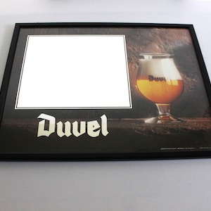 Puede incluir: Un letrero rectangular con marco negro que presenta la palabra Duvel en letras góticas blancas. El diseño incluye una imagen fotográfica de un vaso lleno de cerveza dorada y un área cuadrada blanca grande para exhibición sobre un fondo oscuro.