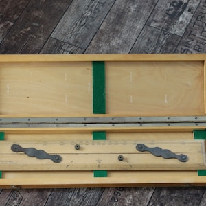 Puede incluir: Una caja de madera con tapa con bisagras, que contiene una herramienta de dibujo con una regla y dos brazos ajustables. La caja está forrada con fieltro verde.