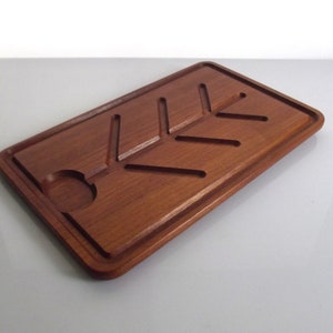 Op de afbeelding: Een rechthoekige houten snijplank met een uitgesneden bladmotief en een cirkelvormige inkeping. De plank is gemaakt van een rijk, roodbruin hout en heeft een opstaande rand. De plank is ongeveer 30 cm lang en 20 cm breed.