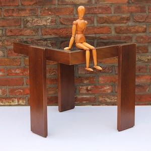 May include: A small, square wooden table with a dark finish. The table has a glass top and four sturdy legs. A wooden artist's mannequin is perched on the table's edge. The table is set against a brick wall.