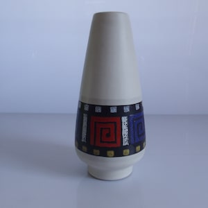 May include: A white ceramic vase with a geometric design in blue, red, and black. The vase has a conical shape and a wide base.