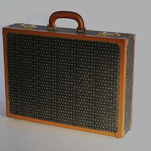 Op de afbeelding: Een vintage koffer met een donkergrijze patroon buitenkant en bruine leren bekleding. De koffer heeft een bruin handvat en goudkleurige sluitingen. Het patroon op de koffer lijkt een herhalend tekstontwerp te zijn.