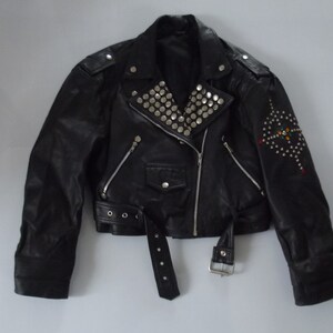 May include: Black leather jacket with silver studs on the collar and shoulder epaulets. The jacket has a diagonal zipper, a belt, and a decorative design of studs and colorful accents on the sleeve.