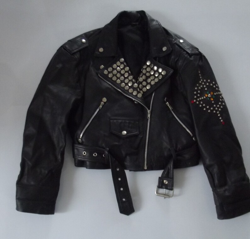 May include: Black leather jacket with silver studs on the collar and shoulder epaulets. The jacket has a diagonal zipper, a belt, and a decorative design of studs and colorful accents on the sleeve.