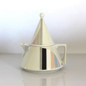 Può includere: Una teiera in ceramica bianca con un design geometrico in tonalità rosa, blu e nero. La teiera ha un coperchio conico e un beccuccio laterale.