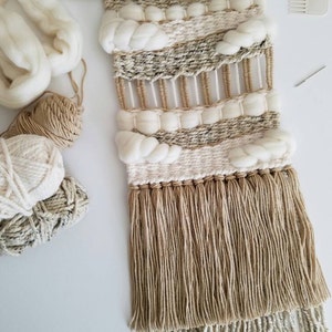 May include: A beige and white woven wall hanging with a wooden dowel at the top and fringe at the bottom. The hanging features a variety of textures and patterns, including woven stripes, pom poms, and knots.
