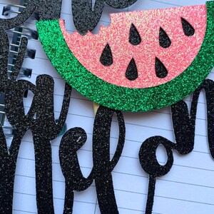 Op de afbeelding: Een glitterende taarttopper met watermeloen thema met het woord "one" en het woord "melon" in zwarte glitter. De watermeloen schijf is roze met zwarte zaden en een groene schil.