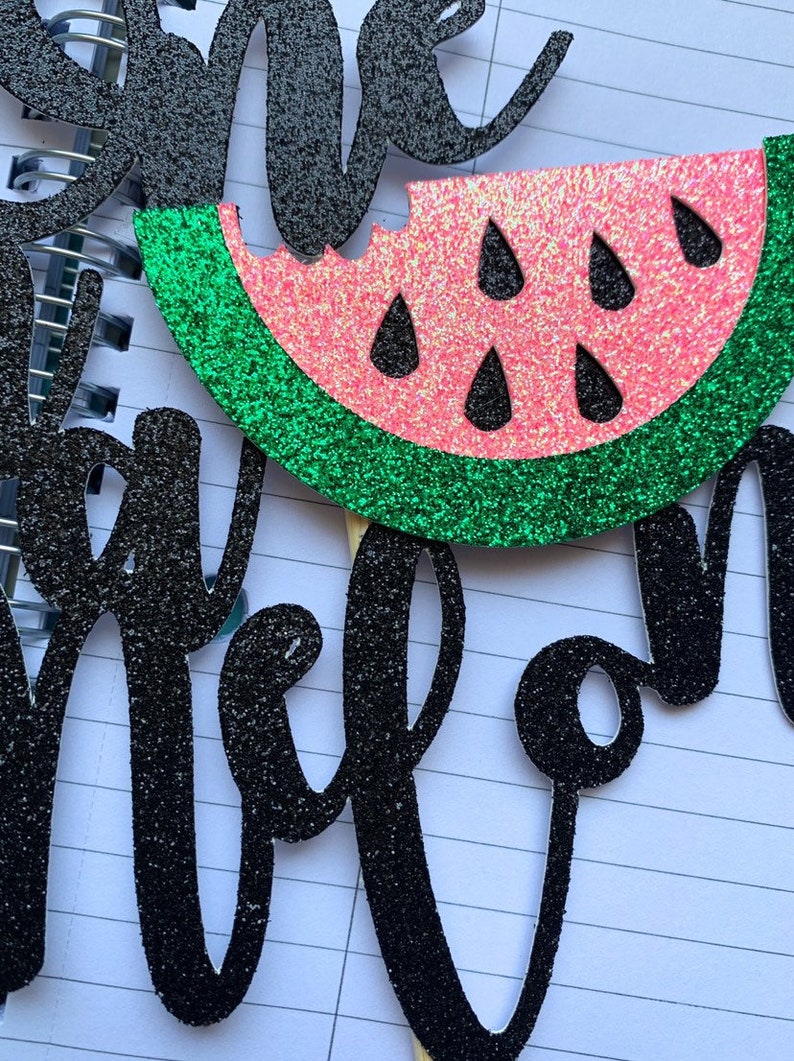 Op de afbeelding: Een glitterende taarttopper met watermeloen thema met het woord "one" en het woord "melon" in zwarte glitter. De watermeloen schijf is roze met zwarte zaden en een groene schil.