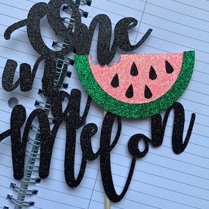 Op de afbeelding: Een zwarte glitter taarttopper met de woorden "one" en "one in a melon". De topper toont een watermeloen met een roze binnenkant, groene schil en zwarte zaden, bevestigd op een houten stokje.