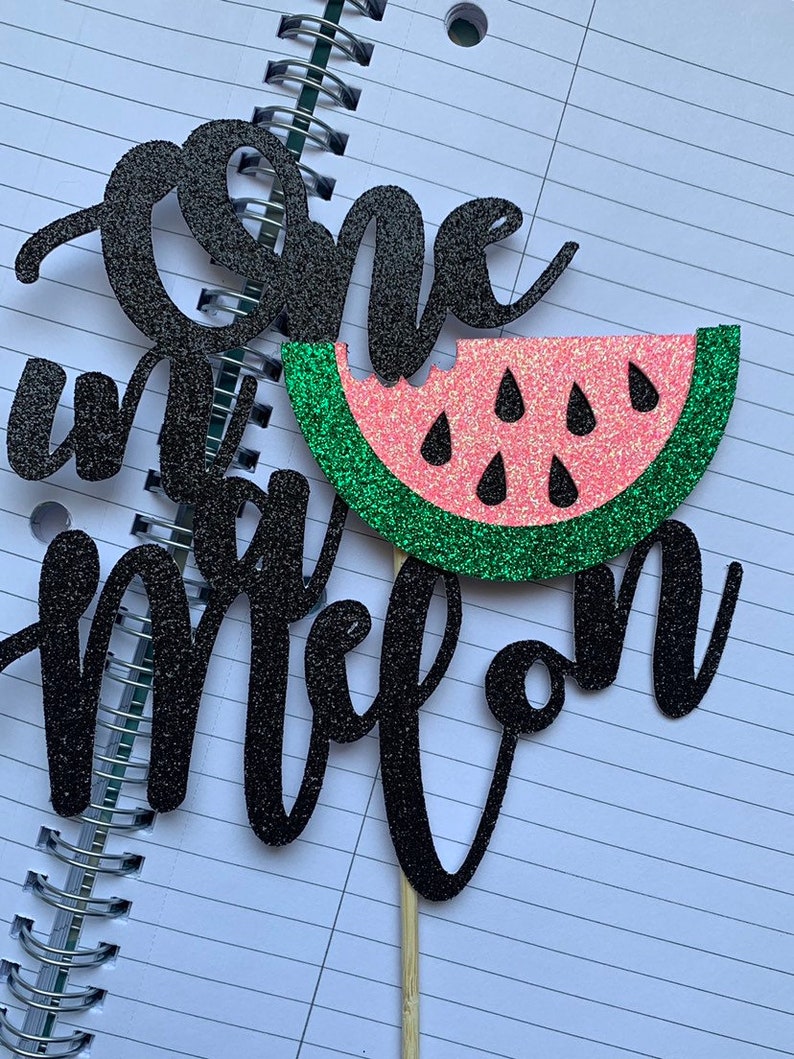 Op de afbeelding: Een zwarte glitter taarttopper met de woorden "one" en "one in a melon". De topper toont een watermeloen met een roze binnenkant, groene schil en zwarte zaden, bevestigd op een houten stokje.