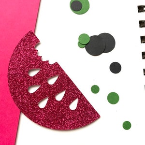 Op de afbeelding: Een glinsterende rode watermeloen schijf met een hap eruit, samen met groene en zwarte cirkelvormige vormen, mogelijk zaden en ogen. Het ontwerp staat op een witte achtergrond met een roze papieren rand.