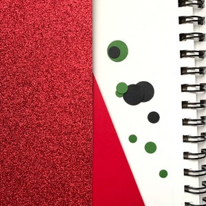 Op de afbeelding: Een spiraalgebonden notitieboek met een rode glitteromslag en een wit interieur. Een rode driehoek en verschillende zwarte en groene cirkelvormige uitsparingen bevinden zich op de witte pagina. De metalen spiraalbinding van het notitieboek is zichtbaar.
