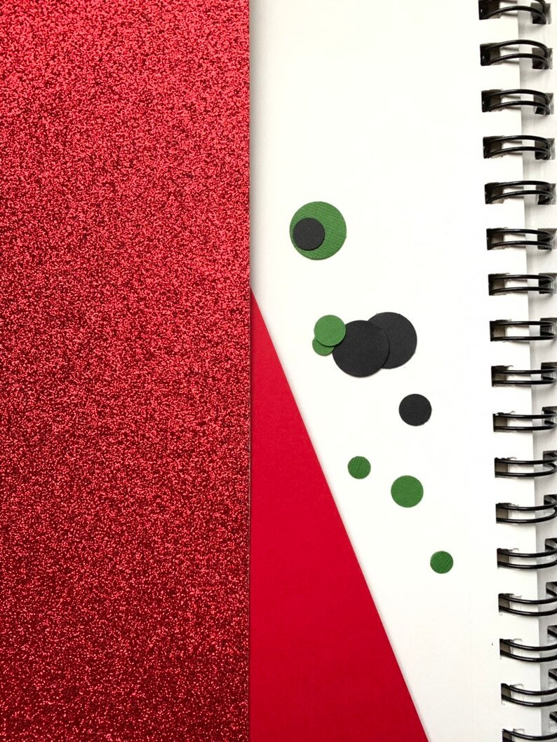 Op de afbeelding: Een spiraalgebonden notitieboek met een rode glitteromslag en een wit interieur. Een rode driehoek en verschillende zwarte en groene cirkelvormige uitsparingen bevinden zich op de witte pagina. De metalen spiraalbinding van het notitieboek is zichtbaar.