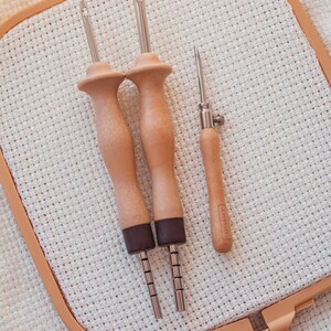 Pu&ograve; includere: Tre aghi da ricamo in legno con punte in metallo e un telaio da ricamo in legno con un tessuto beige teso all'interno.