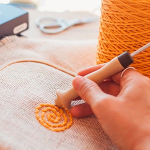 Pu&ograve; includere: Una mano utilizza uno strumento di legno per ricamare un motivo a spirale giallo su un tessuto beige teso in un telaio da ricamo. Un rocchetto di filo giallo si trova nelle vicinanze.
